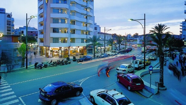 A multi-story hotel with balconies, a street view with cars and a pedestrian crossing, and palm trees lining the sidewalk. - Apartments very close to the beach. Ref.LLORET SUN-24 (Lloret de Mar)