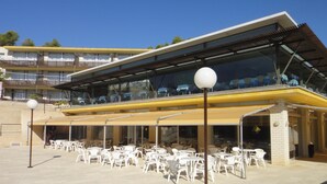 A modern building with a glass facade, outdoor seating area with white plastic chairs and tables, and a yellow awning.