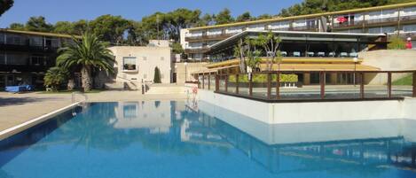 A swimming pool with a clear blue water surface, surrounded by a building with multiple balconies and a covered area with seating.