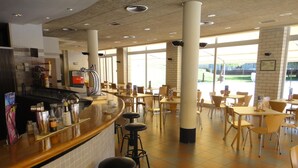 Een ruim café met een bar, meerdere tafels en stoelen, en grote ramen voor natuurlijk lichtinval.