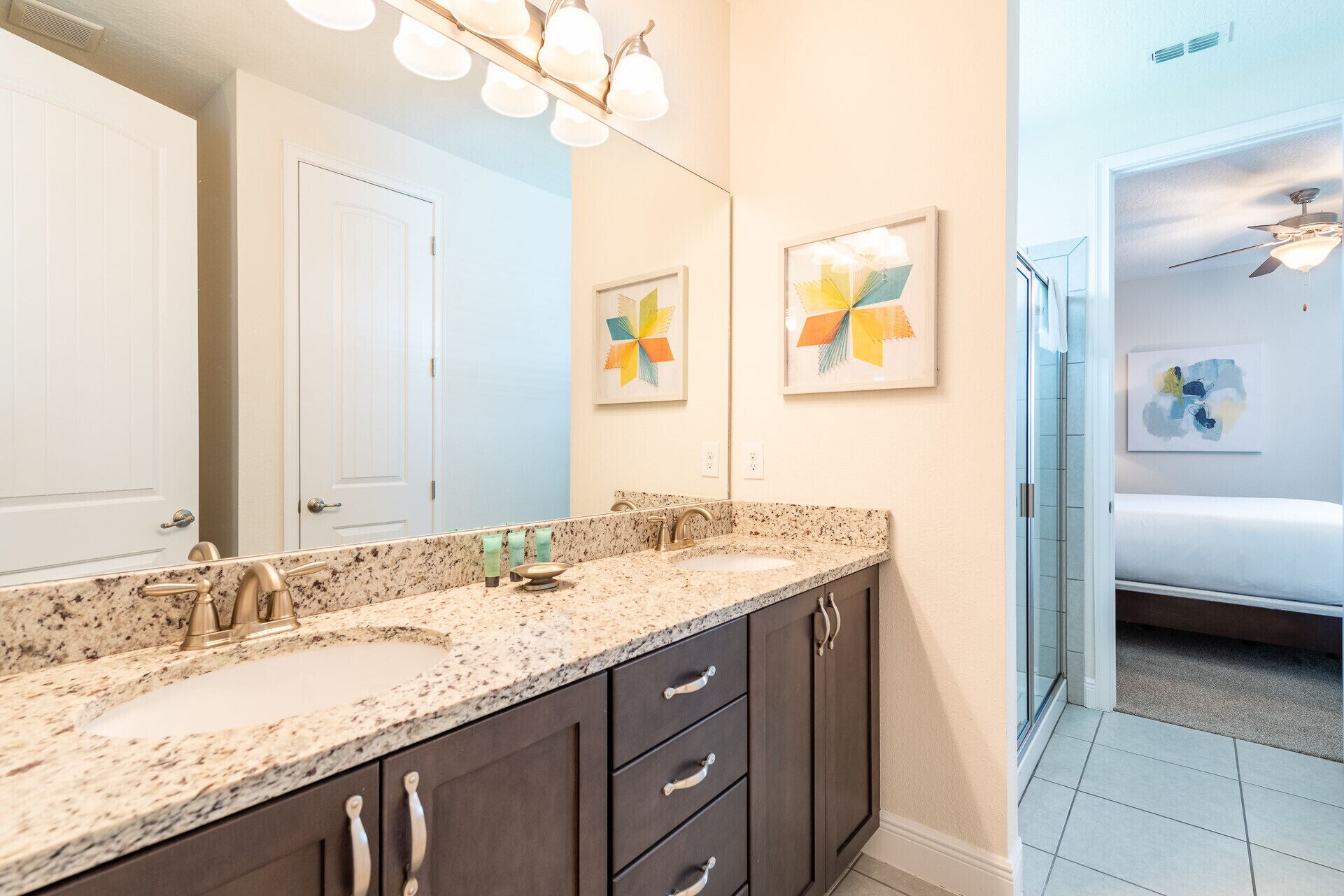 A bathroom with a granite countertop, a double sink, dark cabinets, and a large mirror. There are two wall-mounted light fixtures and two framed artworks above the mirror.
