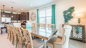 A modern kitchen with dark cabinets, a large window with teal curtains, and a dining area with a glass table and wooden chairs.