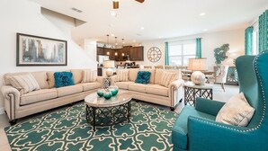 A spacious living room with a sectional sofa, a teal armchair, and a coffee table. A kitchen is visible in the background, and there is a staircase leading to another level.
