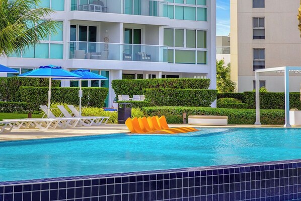 A swimming pool with lounge chairs and umbrellas, surrounded by well-maintained hedges and a modern building with balconies.