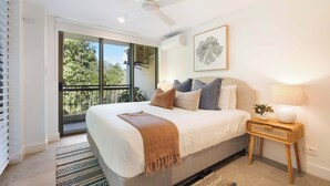 A bedroom with a large bed, a ceiling fan, a window with a view of trees, and a bedside table with a lamp.