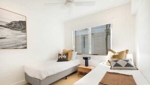 A hotel room with two beds, a small table, a nightstand, a ceiling fan, a window with blinds, and a framed picture on the wall.