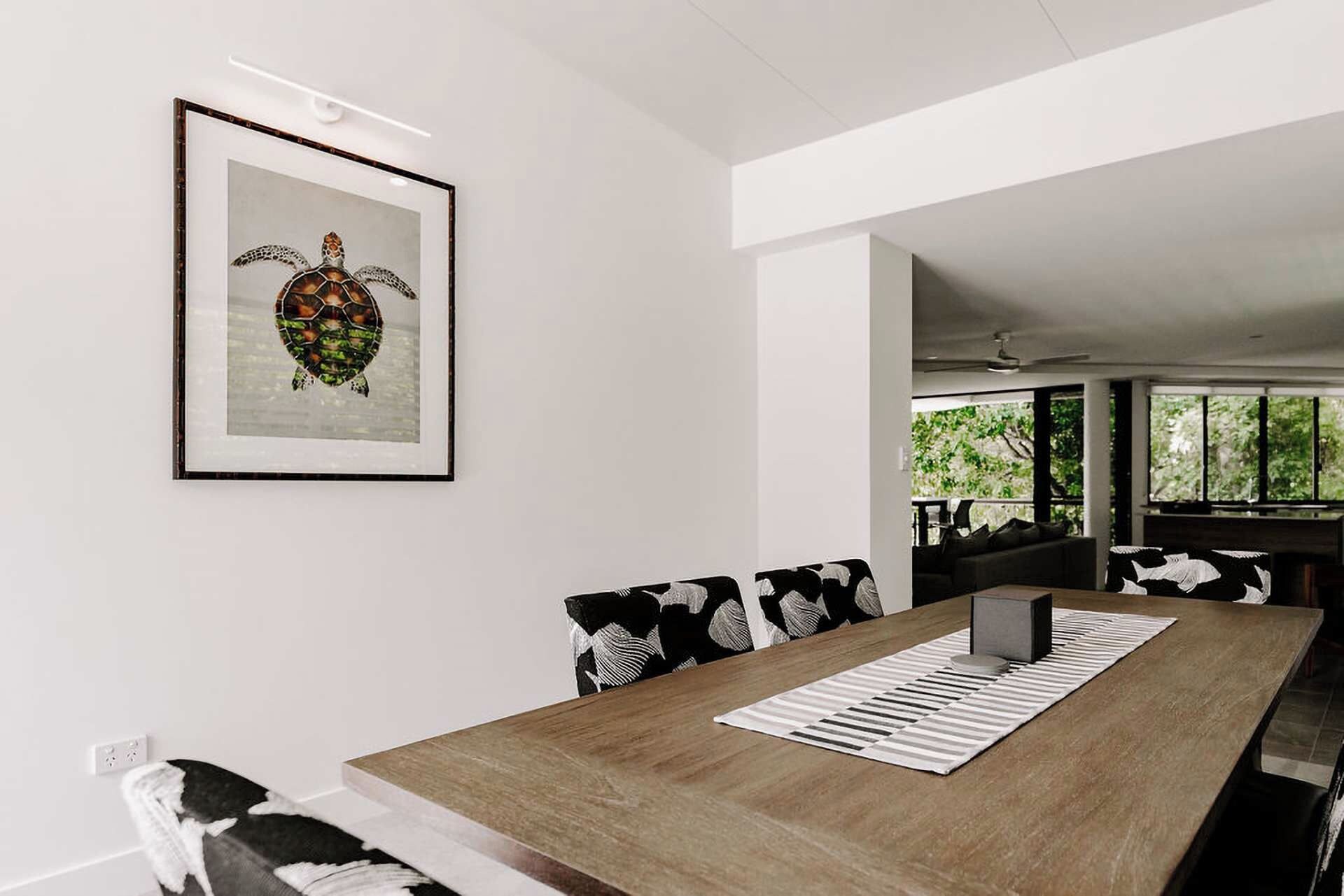 Un espace repas moderne avec une table en bois, des chaises à motifs noir et blanc et une œuvre d’art encadrée représentant une tortue, accrochée au mur.