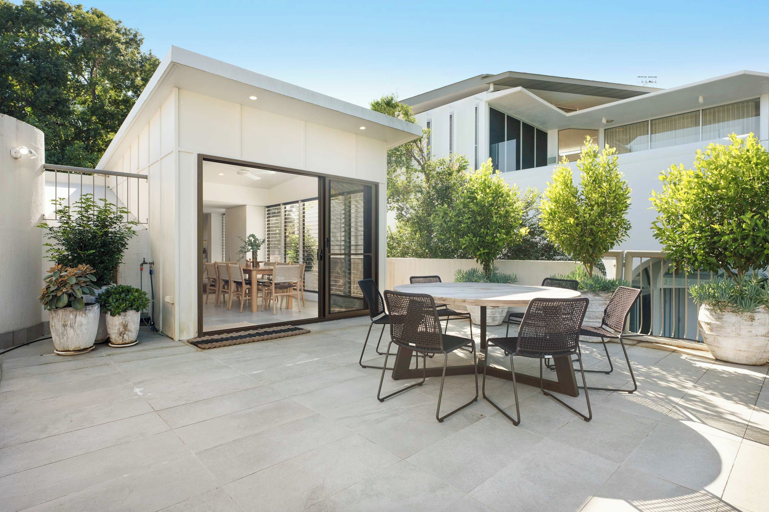 A modern outdoor patio with a round table and chairs, surrounded by potted plants and a glass door leading to an interior space.