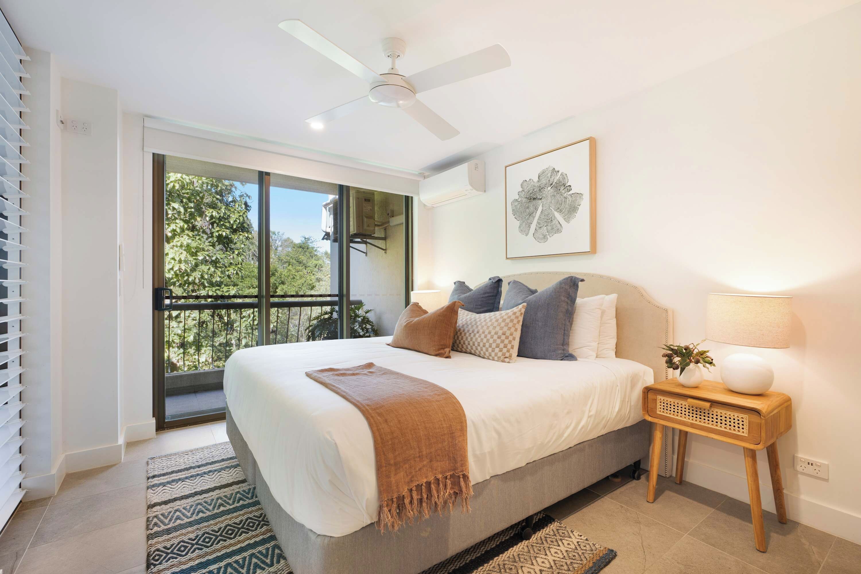 A bedroom with a bed, a ceiling fan, a window with a view of trees, a bedside table with a lamp, and a framed picture on the wall.