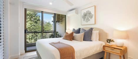 Un dormitorio con cama, ventilador de techo, ventana con vista a árboles, mesita de noche con lámpara y un cuadro enmarcado en la pared.
