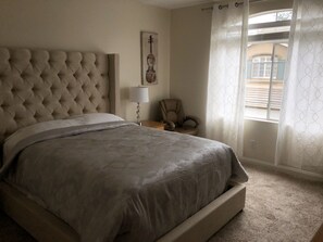 Un dormitorio con cabecera capitonada, cama con colcha gris, lámpara de noche, una silla y una ventana con cortinas estampadas.