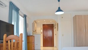 A room with a bed, a wooden table, a television, a kitchenette, and a door leading to another room.