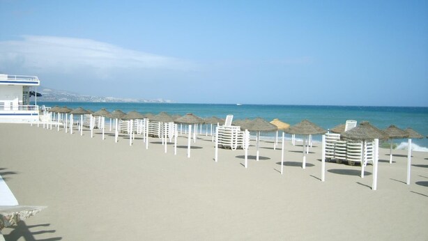 A beach with white umbrellas and lounge chairs, a lifeguard tower, and a clear view of the sea and distant cityscape.
