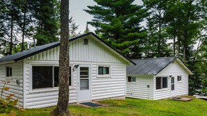 A white log cabin with a large window and a smaller attached structure, surrounded by trees.