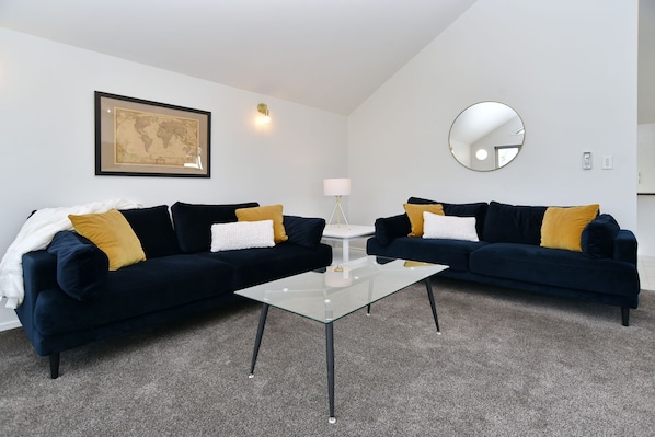 Un salon moderne avec deux canapés bleu foncé, chacun agrémenté d’un plaid blanc et d’un coussin de couleur moutarde. Une table basse en verre est placée au centre, entre les deux canapés. Une carte du monde encadrée est accrochée au mur, au-dessus du canapé situé à gauche.
