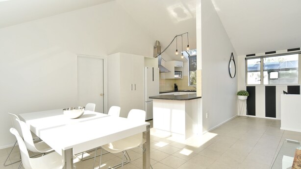 Une cuisine moderne avec des meubles blancs, un coin repas doté d’une table et de chaises blanches, et une grande fenêtre qui laisse entrer la lumière naturelle.