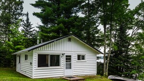 A small, single-story cabin with a gabled roof, a front door, and a large window on the left side. - Wooded Lakeside Cottage ,Fishing, Snowmobiling, Hunting, Boats, Hot Tub (Gwinn)