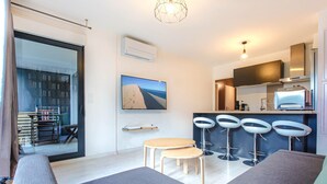 A modern living room with a sofa, a small table, and a TV. There are three bar stools and a kitchen area with a refrigerator and a microwave. - Nice apartment for 4 with parking, near Cité du Vin (Bordeaux)