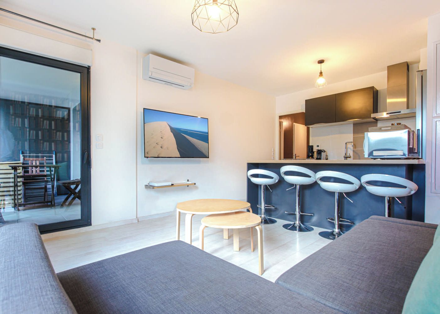 Una sala de estar moderna con un sofá, una mesita y un televisor. Hay tres taburetes de bar y una cocina con un refrigerador y un microondas.
