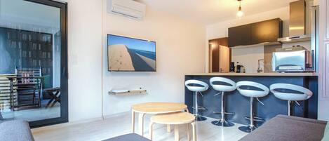 Una sala de estar moderna con un sofá, una mesita y un televisor. Hay tres taburetes de bar y una cocina con un refrigerador y un microondas.