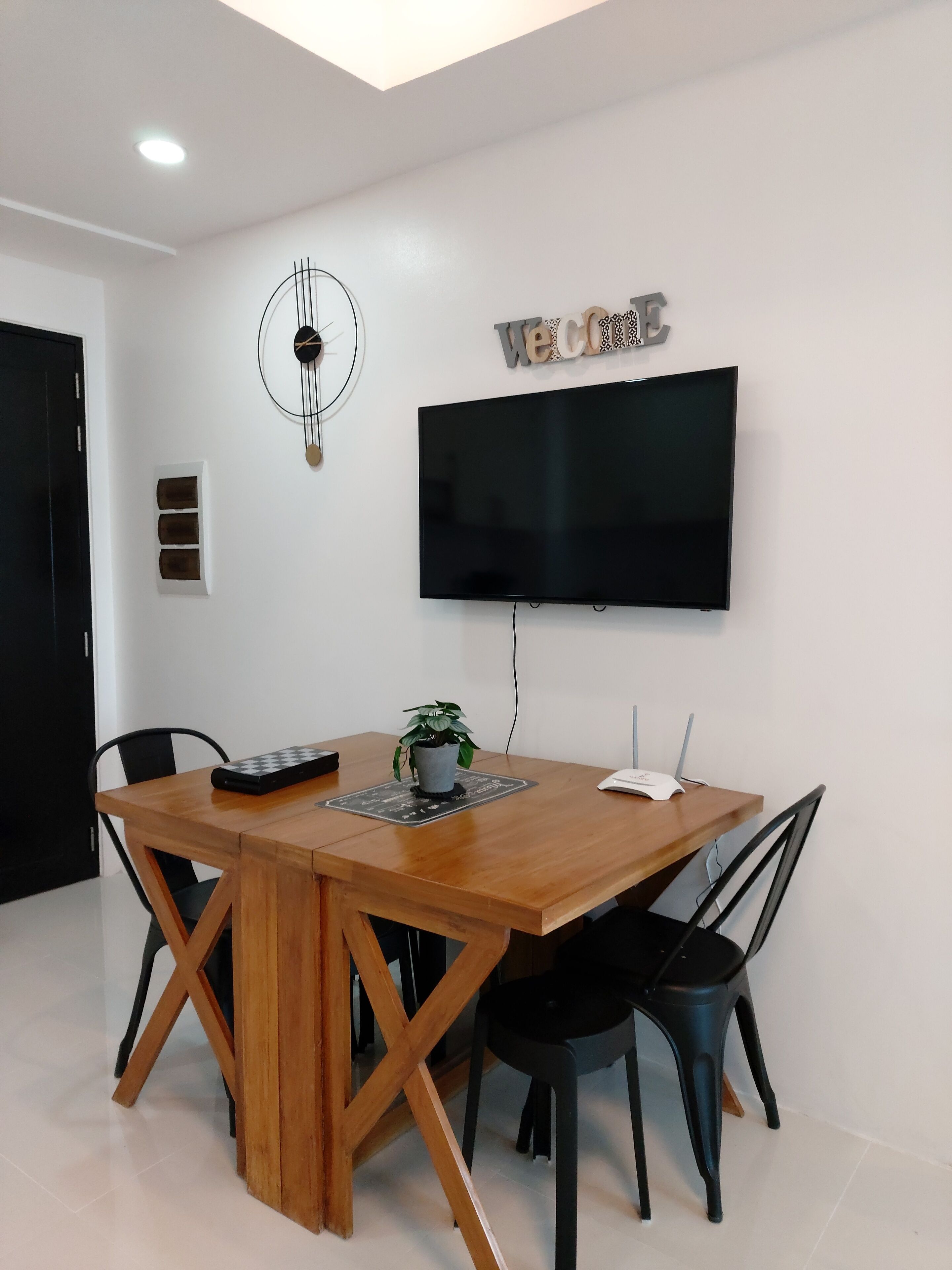 Een moderne eethoek met een houten tafel, zwarte stoelen, een potplant, een tv aan de muur en een decoratieve klok.