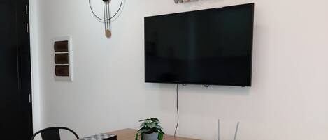 A modern dining area with a wooden table, black chairs, a potted plant, a wall-mounted TV, and a decorative clock.