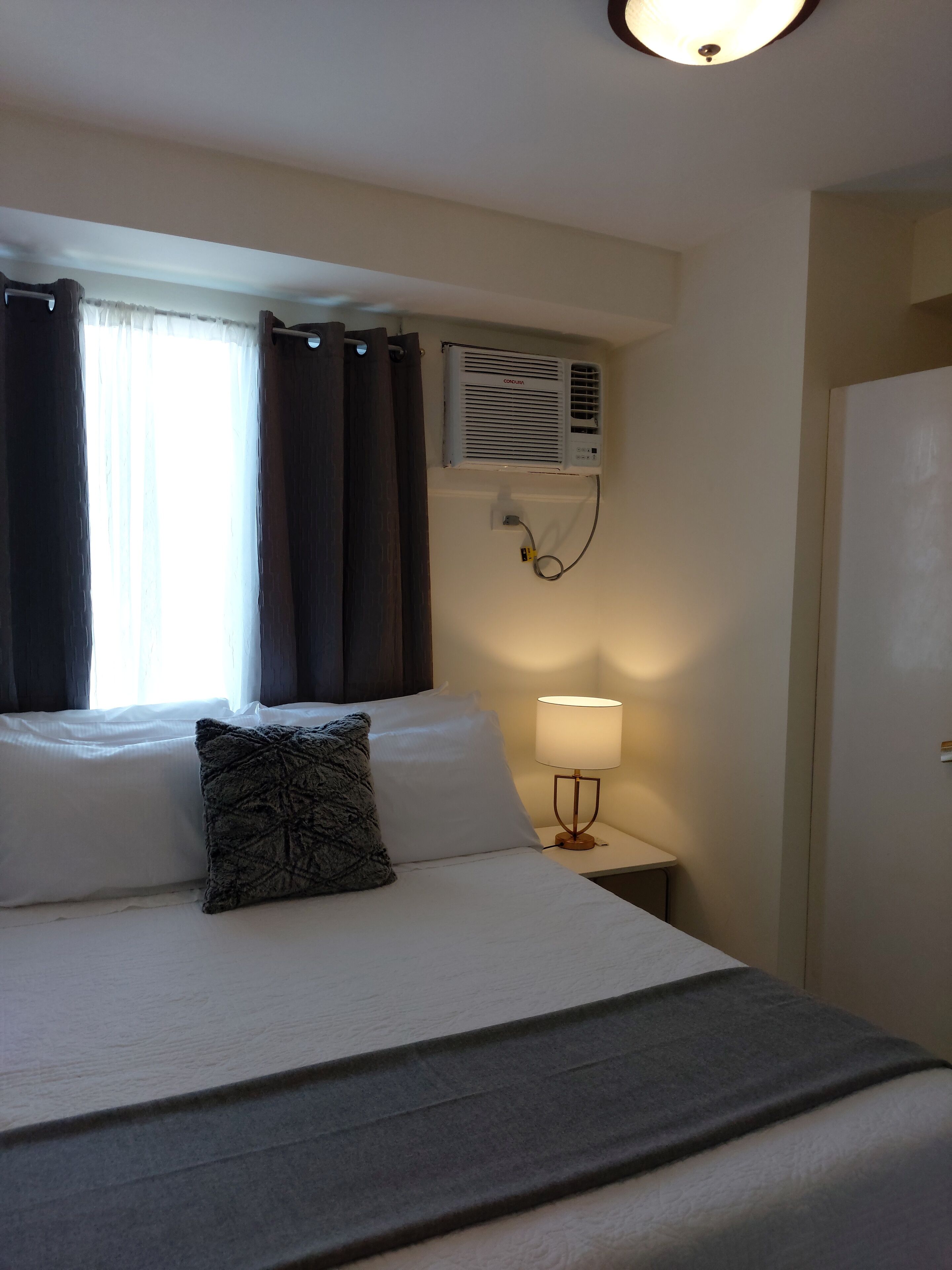 Een hotelkamer met een bed, een bedlamp, een airconditioningsysteem en een raam met gordijnen.