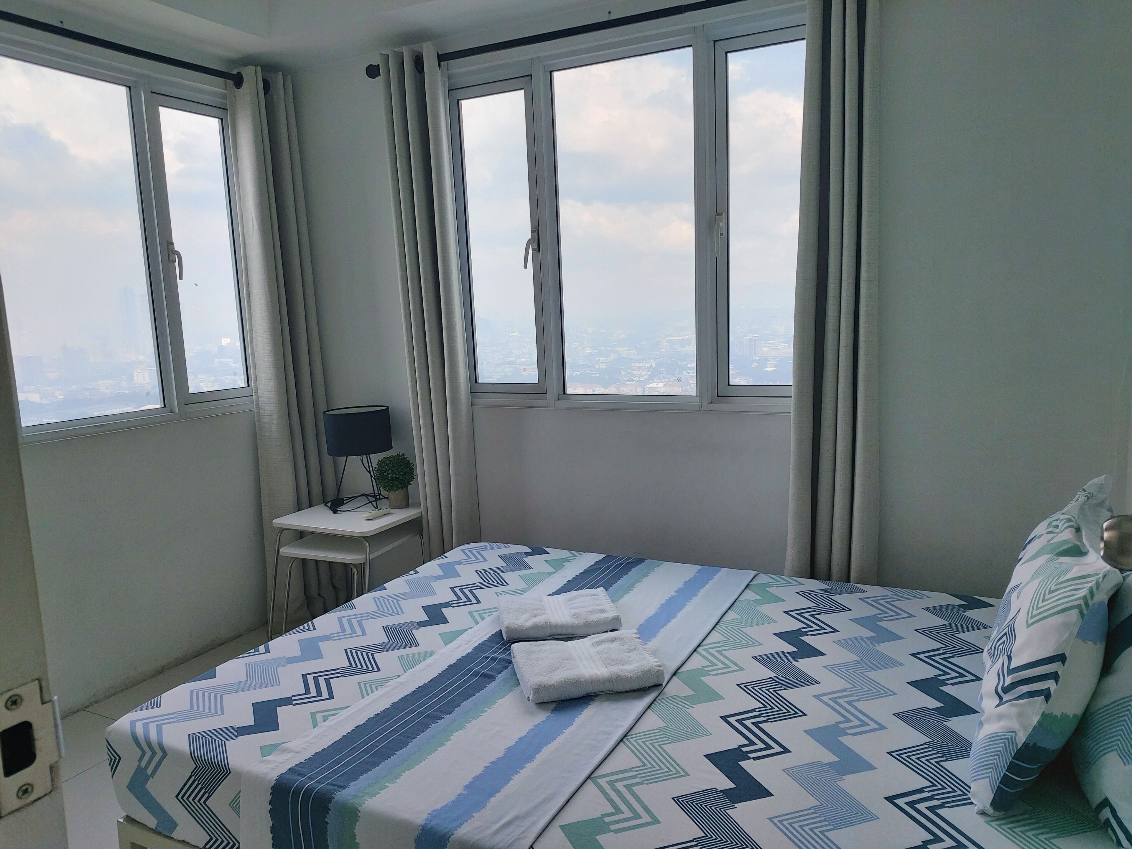 Un dormitorio con cama, dos ventanas, un escritorio con lámpara y vistas a la ciudad.