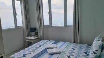 Un dormitorio con cama, dos ventanas, un escritorio con lámpara y vistas a la ciudad.
