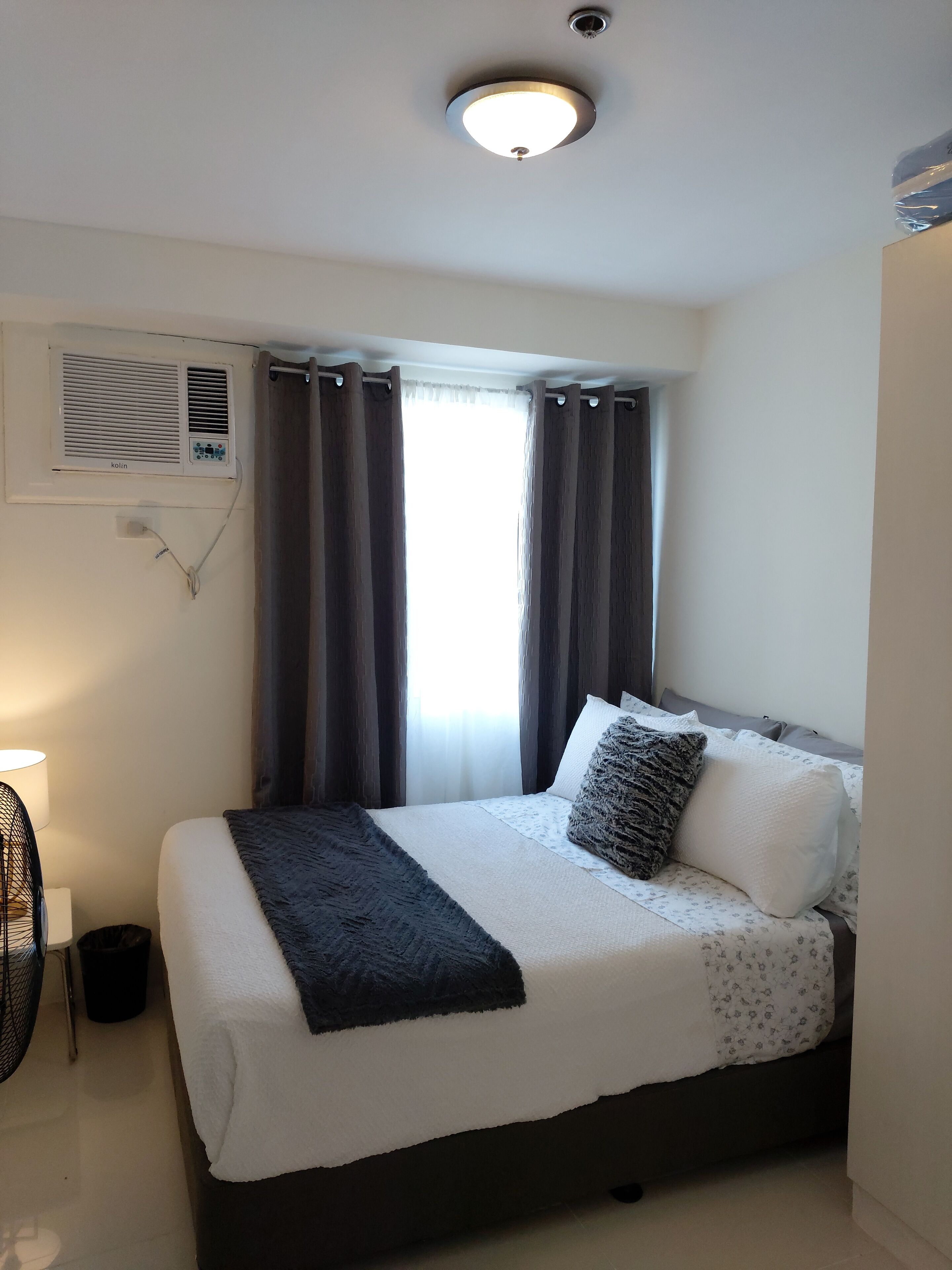 Una cama bien hecha con edredón blanco y gris, un plaid oscuro y varios almohadones, incluyendo uno con textura. Se observa un aire acondicionado instalado en la pared y una ventana con cortinas oscuras.