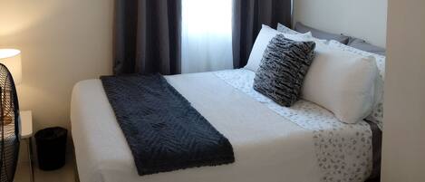 Un lit bien fait, avec une couette blanche et grise, un plaid foncé et plusieurs oreillers, dont un avec une texture particulière. On aperçoit un climatiseur fixé au mur et une fenêtre avec des rideaux foncés.