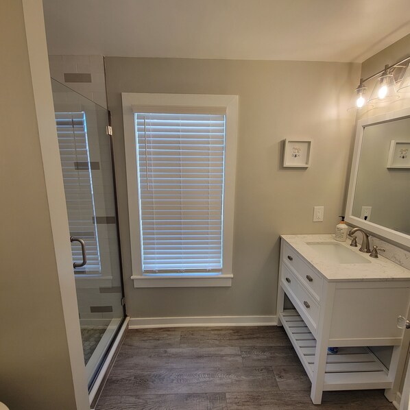 Ein Badezimmer mit bodengleicher Dusche, Waschtisch mit Spiegel und Fenster mit Jalousien.