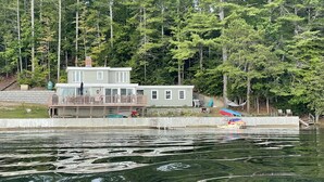 Une maison à deux étages avec un quai qui s’avance sur un lac, entourée d’arbres.