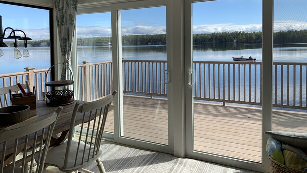 Une pièce avec une table et des chaises en bois, une fenêtre avec des rideaux et une vue sur un lac où se trouve un bateau.