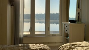 Une chambre avec une grande fenêtre donnant sur un lac paisible, une commode avec un miroir et un lit recouvert d’une couverture à motifs.
