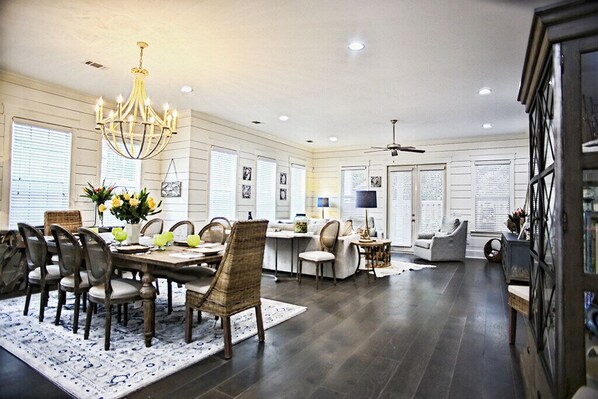 A spacious living room with a dining area, featuring a chandelier, dark wood flooring, and a large window with blinds.