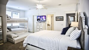 A bedroom with a bunk bed, a single bed, a ceiling fan, a TV, and a door leading to another room. - 1-14 Guest: Pool/Fire Pit/Bikes/Putting Green/Polaris Ranger Included Aug-Dec 25 (St Simons)