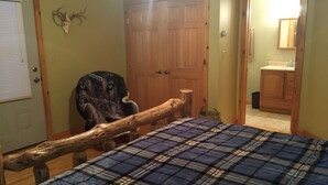 A bedroom with a plaid bedspread, a wooden headboard, a deer head decoration, and a wooden door. - Adirondack Experience (Old Forge)