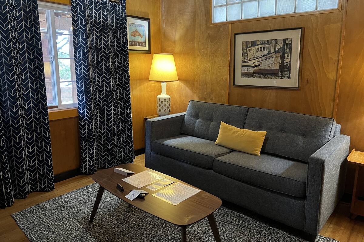 A cozy living room with a grey sofa, a small wooden coffee table, and a lamp. There are two windows with patterned curtains, a framed picture on the wall, and a rug on the floor.