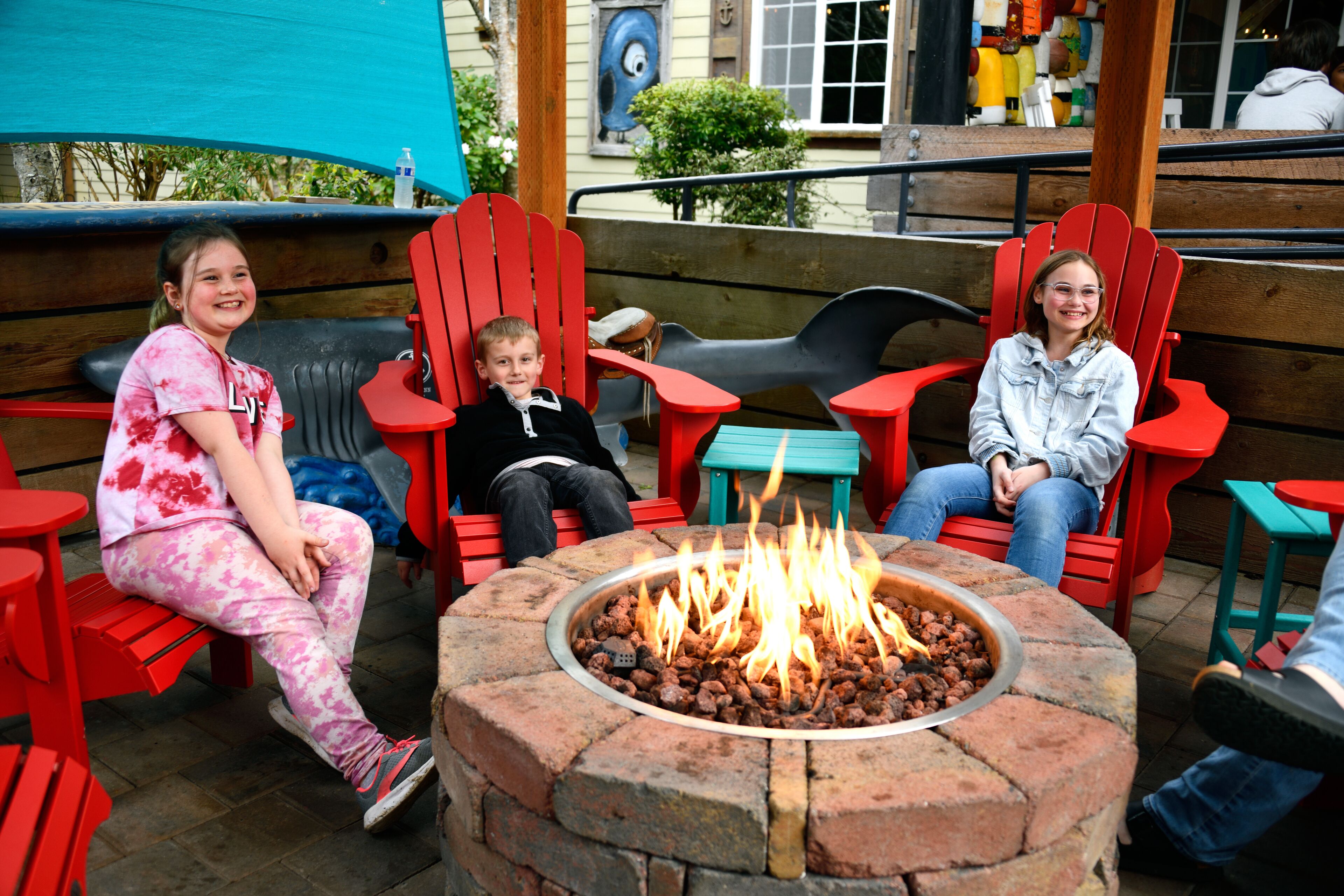 Three children sitting on red Adirondack chairs around a circular brick fire pit.