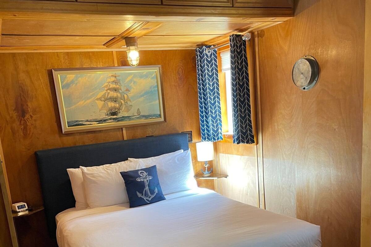A neatly made bed with white linens, a dark headboard, and a single decorative pillow with an anchor design. A painting of a sailing ship is mounted above the bed, and a wall clock is visible to the right.