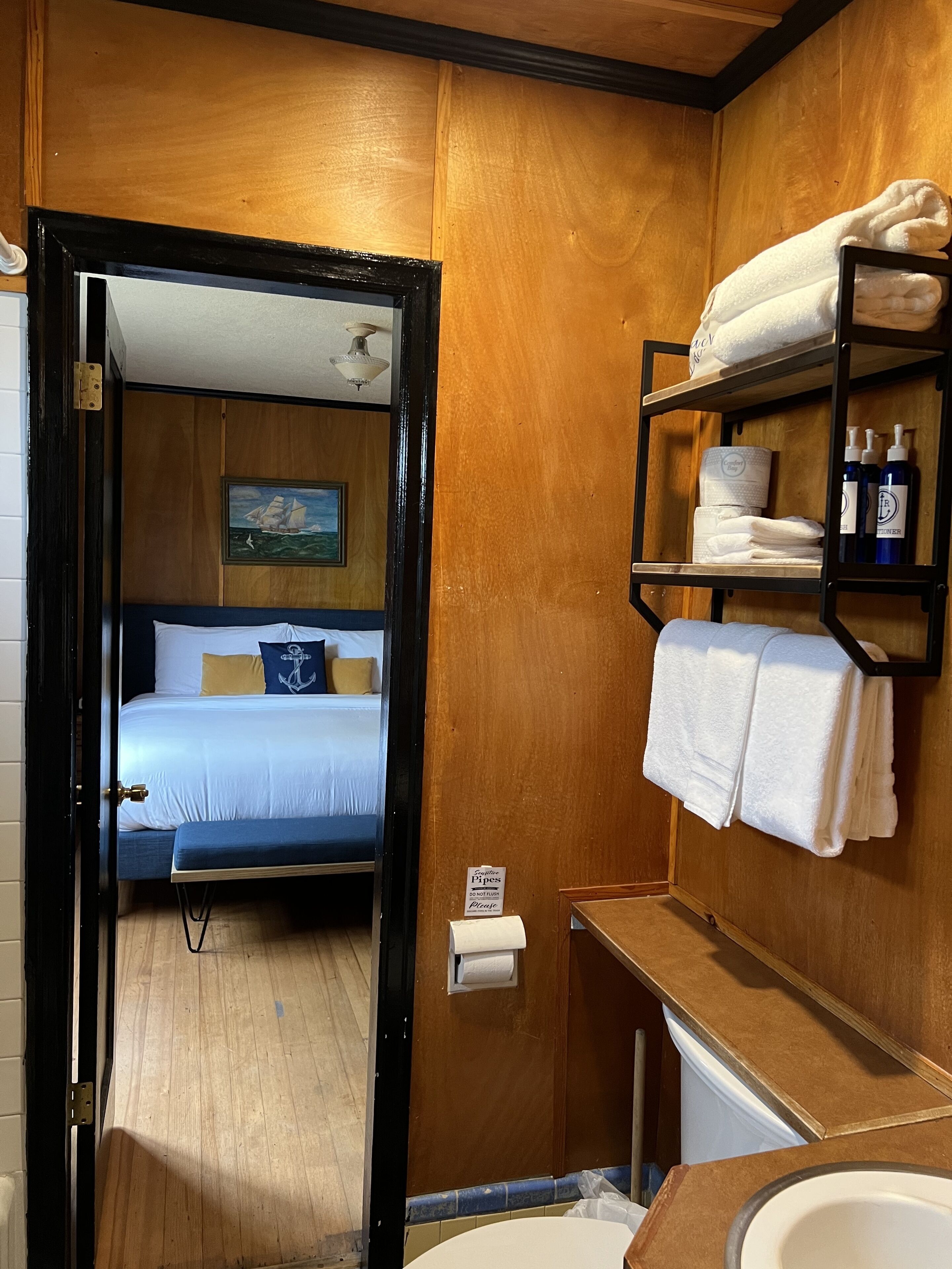 A hotel room with a bed, a wooden wall, a painting, a shelf with towels and toiletries, and a toilet.
