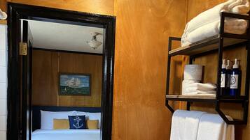 A hotel room with a bed, a wooden wall, a painting, a shelf with towels and toiletries, and a toilet.