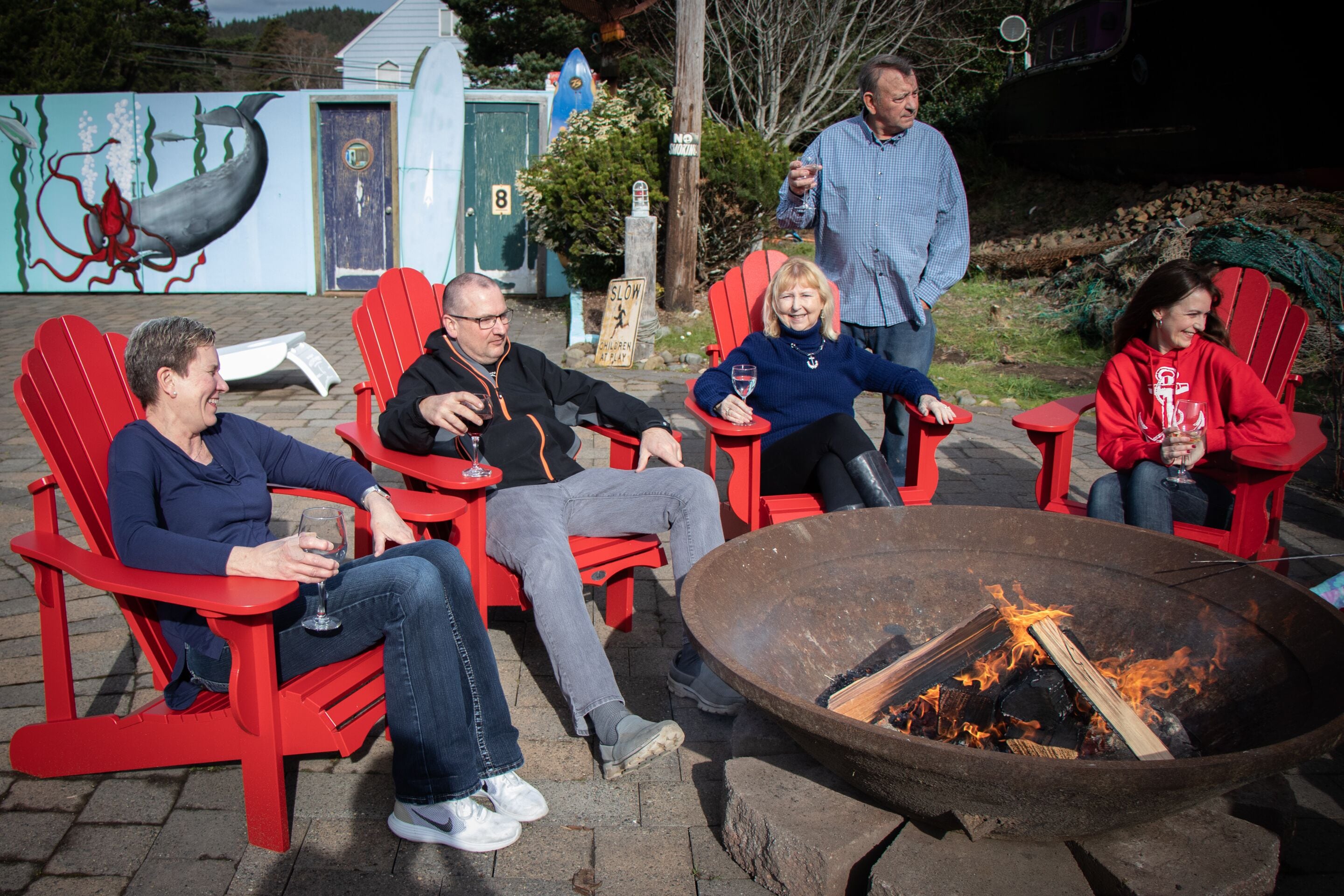 A group of people seated on red Adirondack chairs around a fire pit.