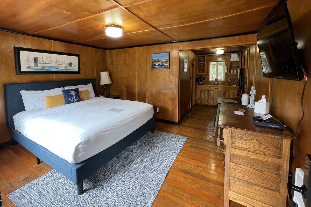 A wooden-paneled room with a bed, a desk, a television, and a kitchen area.