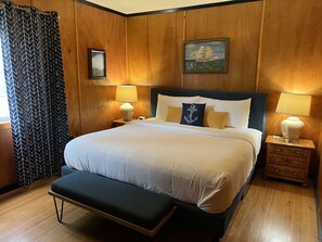 A neatly made bed with white linens, two bedside lamps, a framed picture of a ship, and a wooden nightstand with drawers.