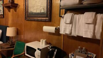 A hotel room with a wooden wall, a framed picture of a ship, a microwave, a chair, a table, and towels on a shelf.