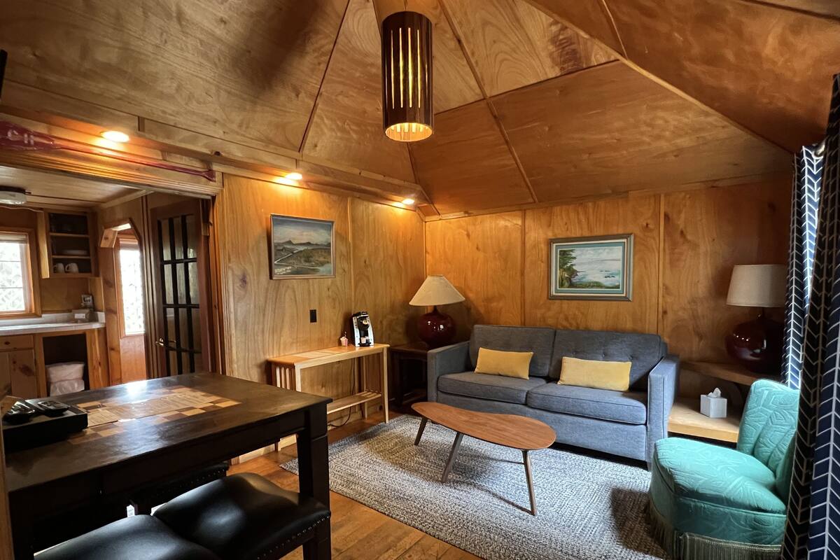 A cozy cabin interior with a wooden ceiling, a sofa, a coffee table, and a dining area with a table and chairs.