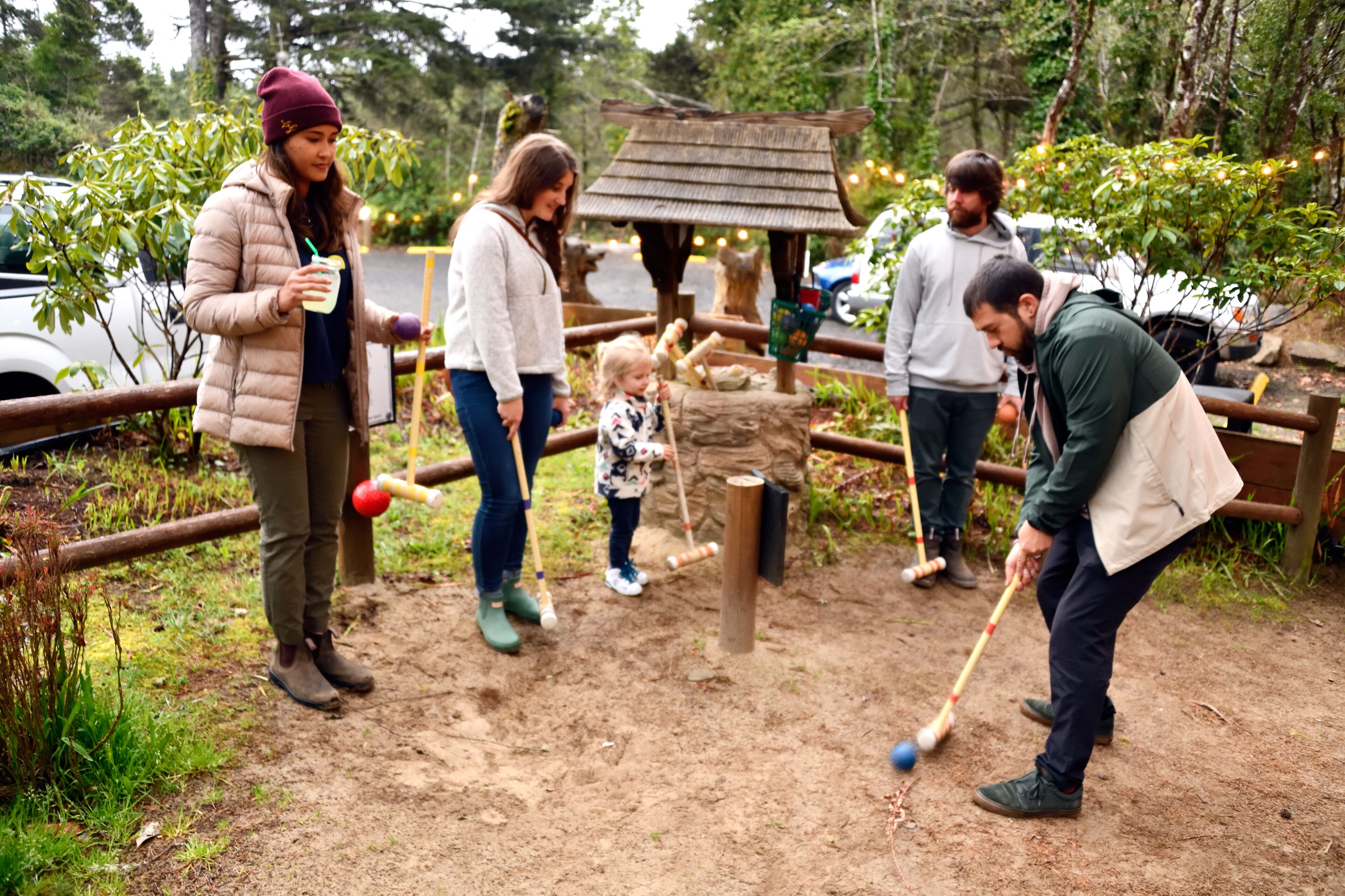 A group of people playing a game with mallets and balls in a park-like setting with wooden structures and greenery.