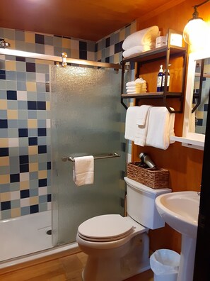 A bathroom with a glass shower enclosure, a toilet, a sink, and shelves with towels and toiletries.
